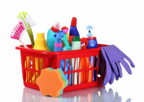 A red plastic basket filled with various cleaning and household supplies, including spray bottles, disinfectants, and scrub brushes, placed on a white surface. The background is plain and neutral, emphasizing the brightly coloured containers and tools. To the right of the basket, a pair of purple rubber cleaning gloves hangs over the side. A colourful, flower-shaped sponge rests at the base of the basket, while the cleaning products feature stickers and caps in shades of yellow, blue, red, and green, with textures that suggest plastic and rubber materials. The overall scene appears to be indoors or on a clean, flat surface suitable for domestic or commercial cleaning tasks, aligning with waste management and rubbish removal services in the local area. The image conveys the concept of household clutter or waste that could be targeted for removal or recycling by companies such as Commercial Waste Removal Ealing, serving clients around west London.