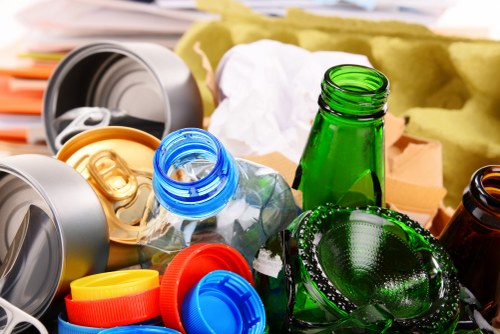 Close-up photograph of a cluttered collection of recyclable waste materials set on a surface, possibly a work area or collection point for rubbish removal. The image prominently features transparent plastic bottles, including a clear bottle with a blue cap, and a green glass bottle positioned centrally. Surrounding these bottles are multiple plastic caps in colors such as red, yellow, and blue, along with crumpled white paper or tissue amid the debris. To the left, part of a grey plastic container or lid is visible, suggesting materials destined for disposal or recycling. The background includes an out-of-focus yellowish surface or furniture, hinting at an indoor or outdoor environment where waste is gathered before collection. The lighting emphasizes the textures of the plastics and glass, highlighting the glossy finishes and the crinkled surfaces of the crumpled paper, consistent with rubbish sorted for removal by a professional waste management service like Commercial Waste Removal Ealing, serving the local area including Ealing in postcode W5.