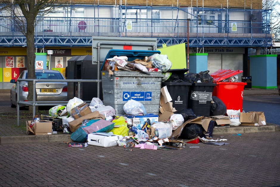 A large accumulation of black plastic rubbish bags piled against a tiled wall in an outdoor urban area, with some bags partially torn, revealing crumpled paper and plastic bottles inside. The bags are